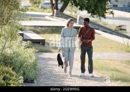 Due giovani uomini d'affari felici in casualwear intelligente che si muovono lungo il parco pubblico e che interagiscono Foto Stock