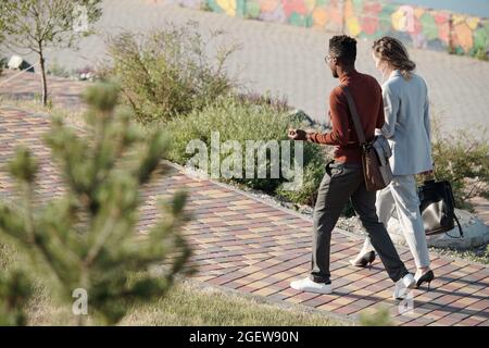 Due giovani colleghi interculturali discutono dei momenti di lavoro mentre si spostano lungo la strada nel parco pubblico Foto Stock