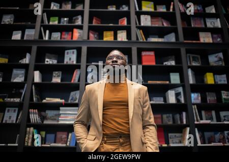 Giovane uomo d'affari elegante in abito e occhiali in piedi contro un grande scaffale in libreria contemporanea Foto Stock