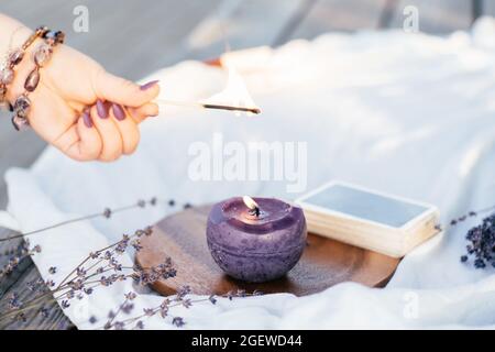 La mano della donna in braccialetto con le unghie lunghe viola accende candela con corrispondenza, vicino al mazzo di carte e fiori di lavanda su superficie bianca e di legno Foto Stock