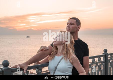 Un uomo e una donna stanno abbracciando il terrapieno e guardano il mare al tramonto. Generazione millenaria. Foto Stock