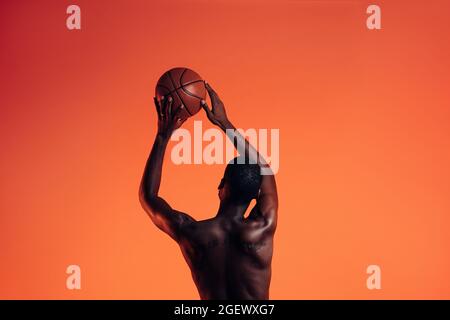 Vista posteriore del giocatore di basket su sfondo arancione. Giovane uomo in studio che si prepara a lanciare la palla del cestino. Foto Stock