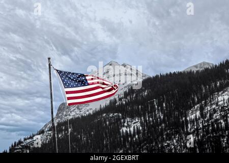 Bandiera americana che sventola contro la montagna scoscesa con neve sotto il cielo coperto. Foto Stock