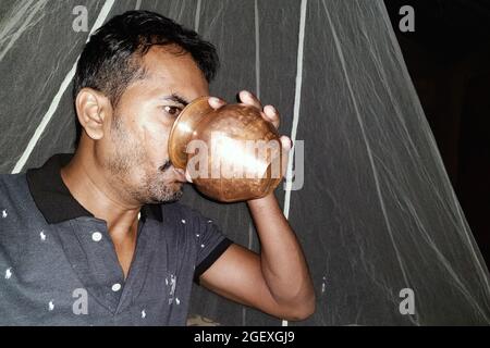 Al mattino, i giovani rurali bevono acqua sui loro letti con una pentola di rame, con zanzariere di fondo Foto Stock