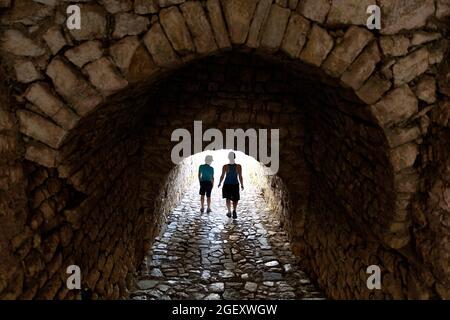 Madre e figlio che camminano attraverso passaggio arco di pietra, vista strada nel villaggio Vuno, con la luce del sole che viene Bebid l'arco, Albania del Sud Foto Stock