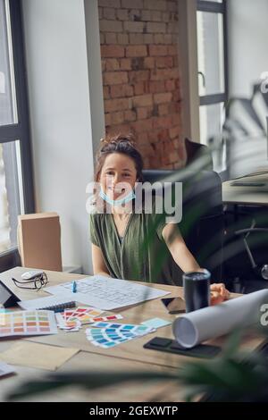 Donna allegra, interior designer con maschera protettiva sorridente alla macchina fotografica mentre lavora con Blueprint e campioni di colore per un nuovo progetto creativo Foto Stock