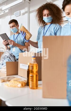 Gruppo di volontari impegnati che indossano uniforme blu, maschere protettive e guanti di calcolo, la selezione di prodotti alimentari donati mentre si lavora in beneficenza Foto Stock