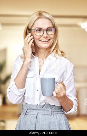 Ritratto di attraente donna d'affari bionda matura sorridente alla macchina fotografica mentre fa la chiamata e beve il caffè, in piedi nel suo ufficio Foto Stock