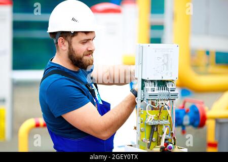 Un ingegnere in un casco e abiti da lavoro regola e ispeziona l'attrezzatura presso lo stabilimento di produzione. Un autentico ritratto di un uomo caucasico con una barba al lavoro. Foto Stock