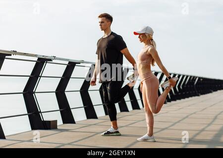 Attraente giovane coppia di fitness facendo esercizi di stretching all'aperto sul mare sul ponte, uomo sportivo e donna che si allenano insieme Foto Stock