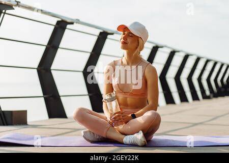 Donna stanca che riposa dopo l'allenamento, sportivo stanco ed esausto seduto sul pavimento su un materassino fitness, con una bottiglia d'acqua all'aperto sul molo Foto Stock