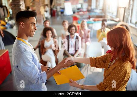 giovane redhead femmina congratularsi con uno studente per aver partecipato con successo al seminario aziendale, dandogli il certificato in cartella. immagine fuoco selettivo. Foto Stock