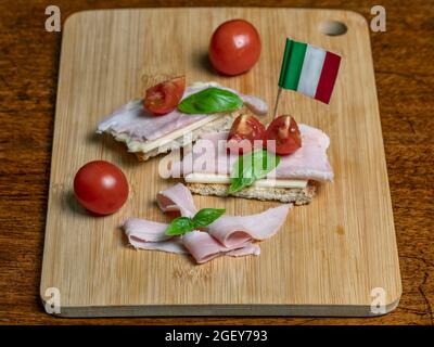 Gustoso spuntino con pane tostato coperto con fette di formaggio e prosciutto, foglie di basilico, pomodori ciliegini su tagliere di legno e bandiera italiana Foto Stock