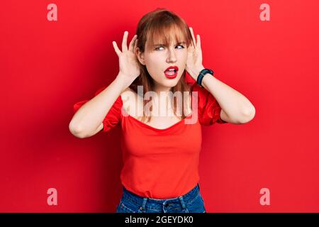 Giovane donna rossa che indossa una t-shirt rossa casual cercando di sentire entrambe le mani sul gesto dell'orecchio, curioso per pettegolezzi. Problema uditivo, sordi Foto Stock