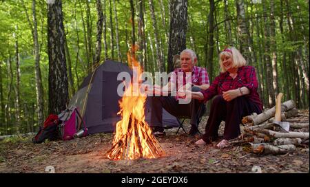 Nonno anziano che cucinano salsicce di frittura sul fuoco in legno al campeggio Foto Stock