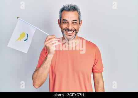 Bell'uomo di mezza età con capelli grigi che reggono la bandiera di cipro guardando positivo e felice in piedi e sorridendo con un sorriso sicuro mostrando i denti Foto Stock