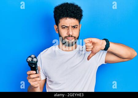 Giovane uomo arabo con barba che tiene la macchina elettrica del rasoio con faccia arrabbiata, segno negativo che mostra dissimile con i pollici giù, concetto di rifiuto Foto Stock