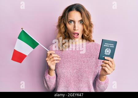 Giovane donna caucasica con bandiera italiana e passaporto in faccia d'urto, scettica e sarcastica, sorpresa dalla bocca aperta Foto Stock