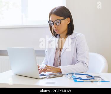 Concentrato afro americano medico donna utilizzando il computer portatile mentre si siede sul posto di lavoro in clinica medica Foto Stock
