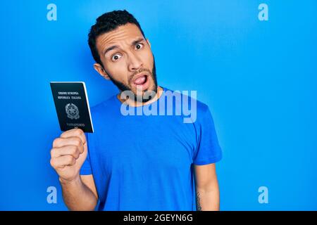 Uomo ispanico con barba che tiene il passaporto italia in faccia shock, che guarda scettico e sarcastico, sorpreso con bocca aperta Foto Stock