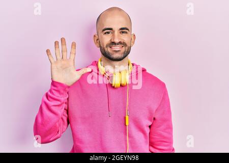 Giovane uomo calvo che indossa abiti da palestra e utilizzando le cuffie mostrando e puntando con le dita numero cinque, sorridendo sicuro e felice. Foto Stock