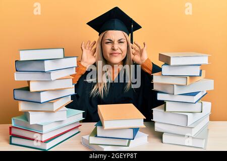 Giovane donna caucasica che indossa un abito da cerimonia di laurea seduto sul tavolo cercando di sentire entrambe le mani sul gesto dell'orecchio, curioso per pettegolezzi Foto Stock