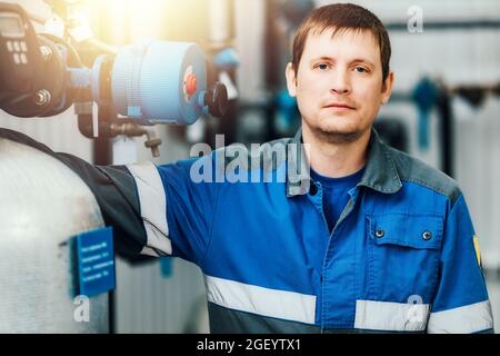 Ritratto di un giovane lavoratore caucasico in tute contro lo sfondo di attrezzature caldaia. Fabbro o operatore locale caldaia a gas. Un vero lavoratore in un impianto di produzione. Foto Stock