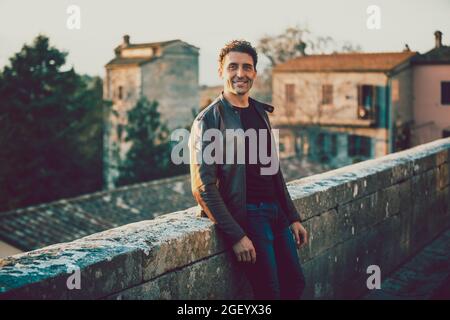 Affascinante uomo sorridente all'aperto con giacca e jeans in pelle. Appoggiati su una parete bassa. Vecchie case di un villaggio sullo sfondo. Moda invernale. Foto Stock