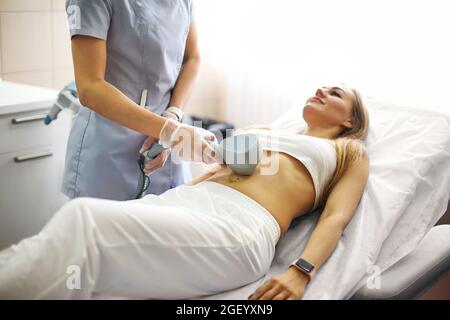 Giovane bella donna bionda sorridente ottenere trattamento professionale di cura della pelle in clinica di bellezza mentre si stende su sedia confortevole, medico utilizzando speciale Foto Stock