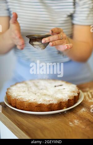 Foto ritagliata di donna in casalinga grembiule cospargere deliziosa torta con zucchero a velo attraverso il filtro, facendo tocchi di finitura sulla mela appena sfornata Foto Stock