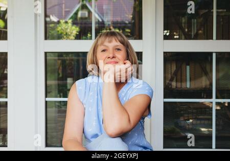 Felice nonna anziana sorridente alla macchina fotografica mentre si siede sulla terrazza della sua casa di campagna e si riposa testa a portata di mano, aspettando i nipoti su di noi Foto Stock
