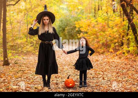 Scatto corto di madre di famiglia e figlia in piedi all'esterno con faccia spaventosa lanterna di zucca tra loro sul percorso pieno di foglie cadute nel parco autunnale, Foto Stock