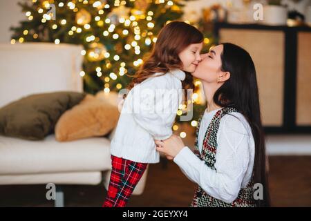 Bella madre amorevole baciare dolcemente la figlia carina contro l'albero di Natale decorato in modo festivo con luci scintillanti sullo sfondo, famiglia felice Foto Stock