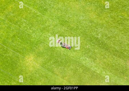 Vista aerea di una piccola figura di uomo lavoratore rifilare erba verde con mashine falciatura sul campo di calcio stadio in estate. Foto Stock