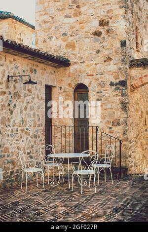 Piccolo tavolo con sedie bianche all'esterno di una casa in mattoni e pietra in un antico villaggio italiano. Scena vintage. Foto Stock