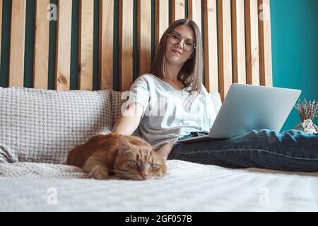 Giovane donna che usa un computer portatile per lavorare, sdraiata sul letto con un gatto in camera da letto, a casa. Orari flessibili e lavoro a distanza Foto Stock