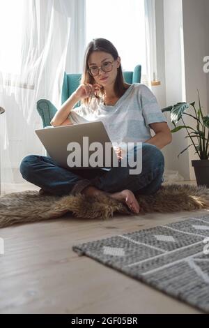 Giovane donna che usa un computer portatile per lavorare, seduto sul pavimento a casa, orari flessibili e lavoro a distanza Foto Stock