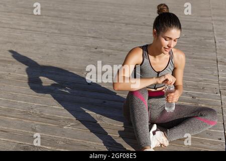 Colpo all'aperto di attraente donna fitness che ha una pausa dopo jogging, seduta sul pavimento e acqua potabile, allenandosi vicino al mare Foto Stock