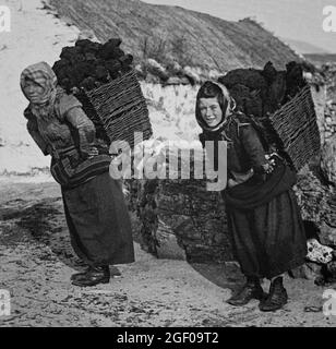 Una scena di inizio 20 ° secolo di una donna che trasportano il tappeto erboso (o torba) in cestini da frusta tradizionali dalla palude al loro cottage di paglia sulla costa occidentale dell'Irlanda. Foto Stock