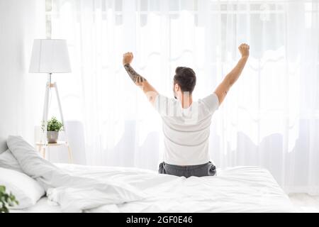 Rilassato giovane uomo che si svegliava a letto e stretching le braccia, di fronte alla finestra, vista posteriore, spazio libero Foto Stock