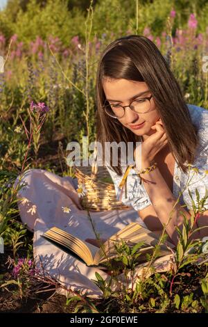 Una giovane ragazza premurosa giace sull'erba verde e legge un libro illuminato dal sole che tramonta. Preparazione di uno studente per l'esame. Lettura carta b Foto Stock