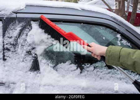 Problemi invernali dei conducenti di auto. Primo piano di una mano di una donna che spazzola la neve dalla sua auto con una spazzola rossa in una fredda giornata invernale nevosa. Foto Stock