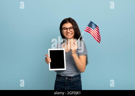 Studiare all'estero online. Studentessa indiana teen femmina che mostra tablet con schermo vuoto, tenendo bandiera americana, mockup Foto Stock