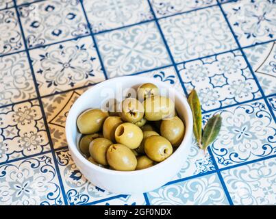 Piccola ciotola bianca con uno spuntino di olive verdi su un tavolo con piastrelle tradizionali blu mediterraneo sotto un ulivo Foto Stock