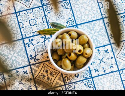 Piccola ciotola bianca con uno spuntino di olive verdi su un tavolo con piastrelle tradizionali blu mediterraneo sotto un ulivo Foto Stock