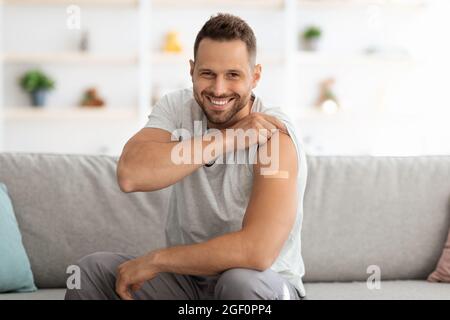 Felice giovane vaccinato uomo mostrando spalla con cerotto medico dopo l'iniezione di vaccino e sorridendo ampiamente a macchina fotografica Foto Stock