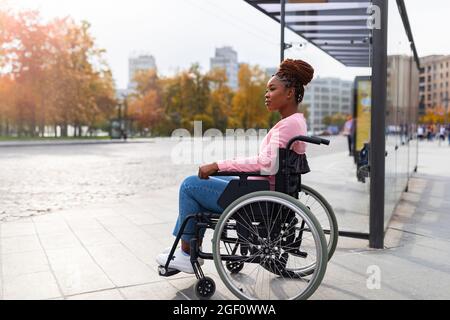 Donna paraplegica nera in sedia a rotelle in attesa sulla fermata dell'autobus, non può salire a bordo di un veicolo adatto a persone disabili Foto Stock