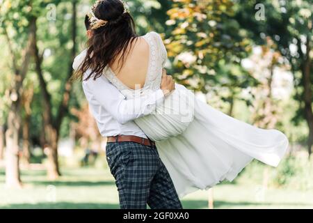 Felice coppia di nozze, lo sposo ammira la sposa felice, tiene la sposa nelle sue braccia e la vortice, nel parco in un giorno di sole estivo, felice matrimonio Foto Stock