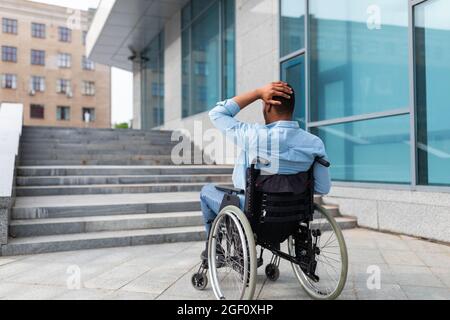 Uomo nero irritato in sedia a rotelle che non ha la possibilità di entrare nell'edificio senza rampa, all'aperto Foto Stock