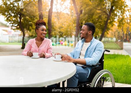 Ragazzo nero disabili in sedia a rotelle che ha appuntamento con la donna in un caffè all'aperto, bere caffè, godere di parlare al parco autunnale Foto Stock